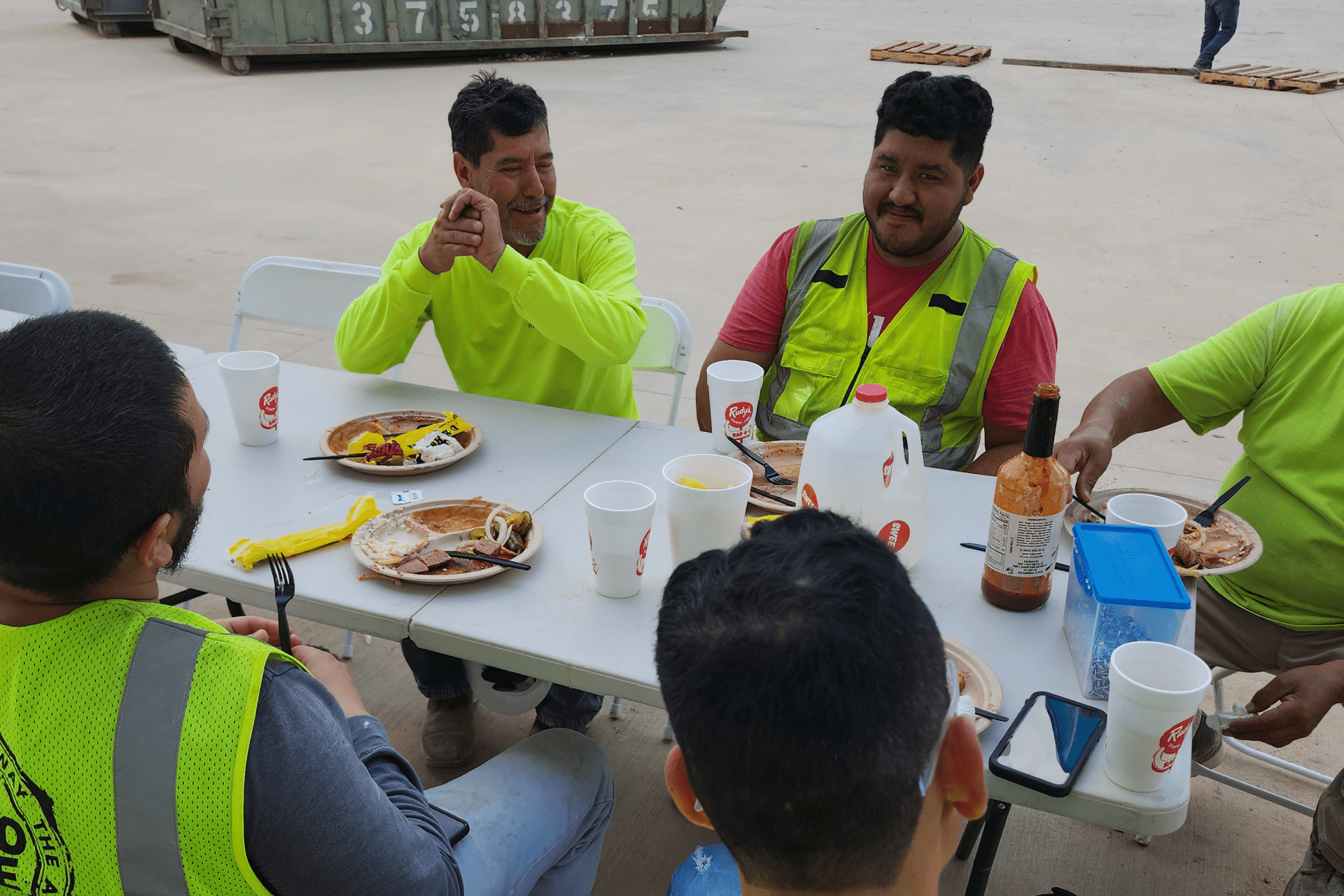 a group of construction workers gathered around a white folding table, enjoying a meal together